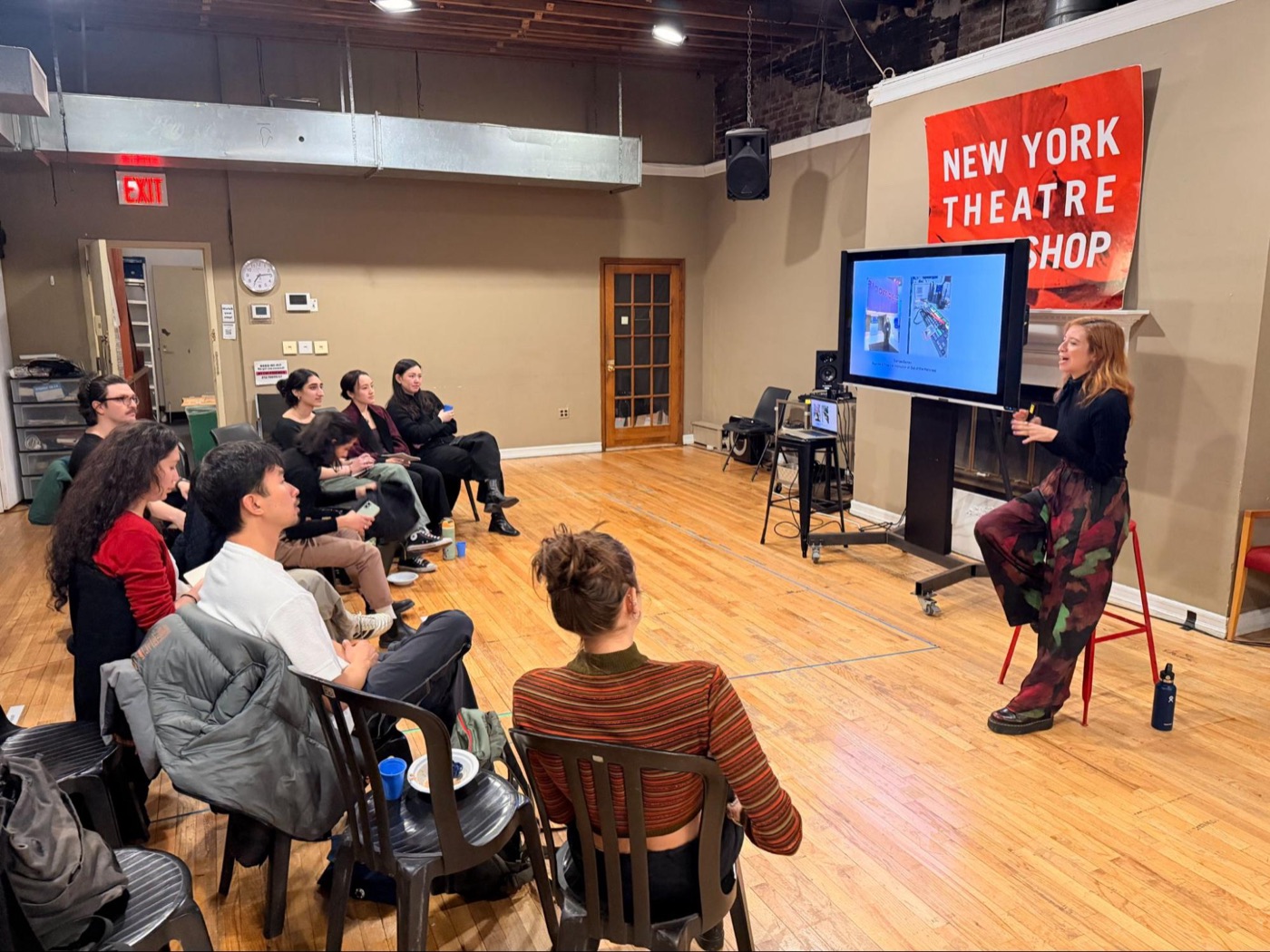 A group of participants seated in rows at an Out of the Mattress workshop at New York Theatre Workshop, facing founder Larissa Gomes, who is presenting, and a screen